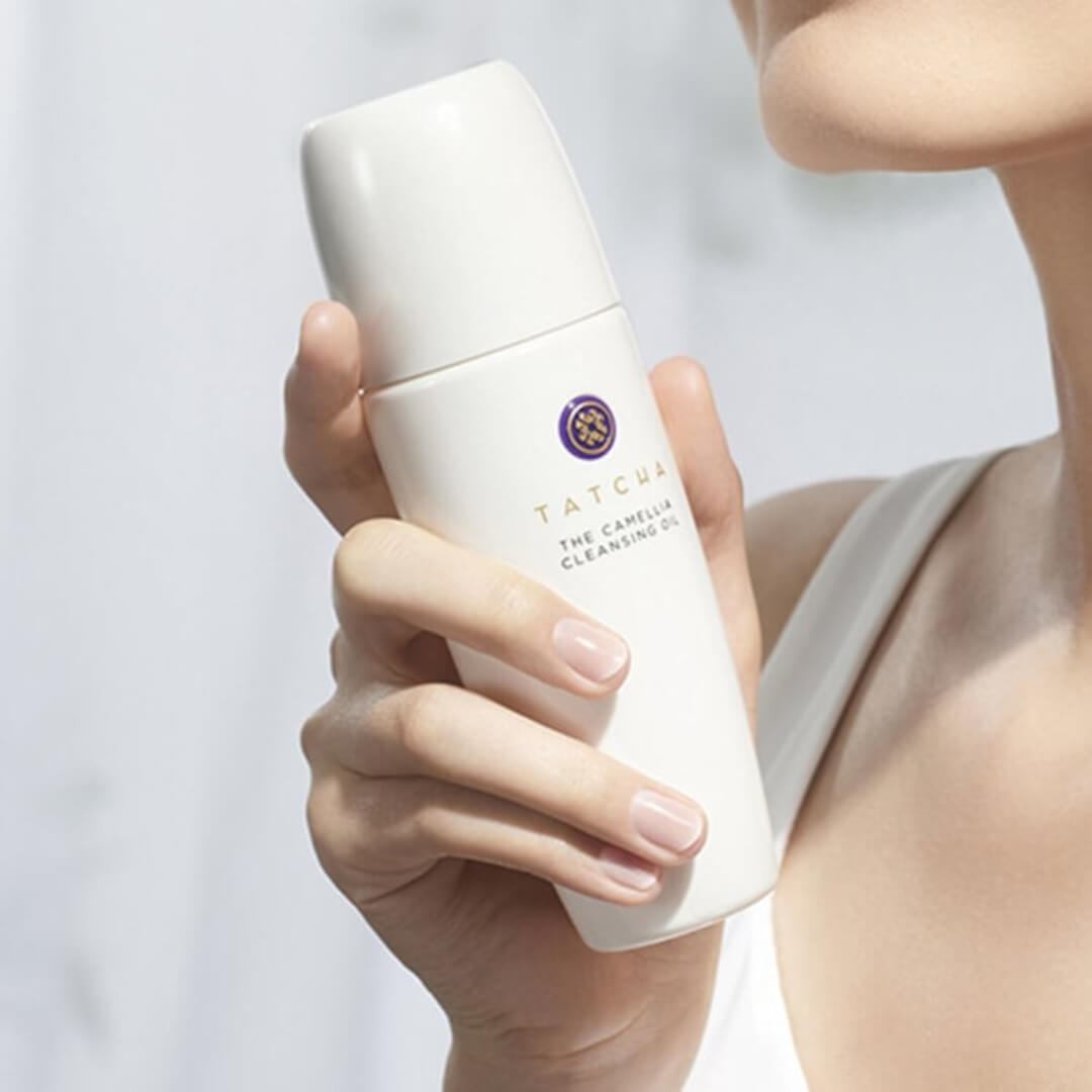 Woman holding Tatcha The Camellia Cleansing Oil, demonstrating use for removing makeup and cleansing skin.